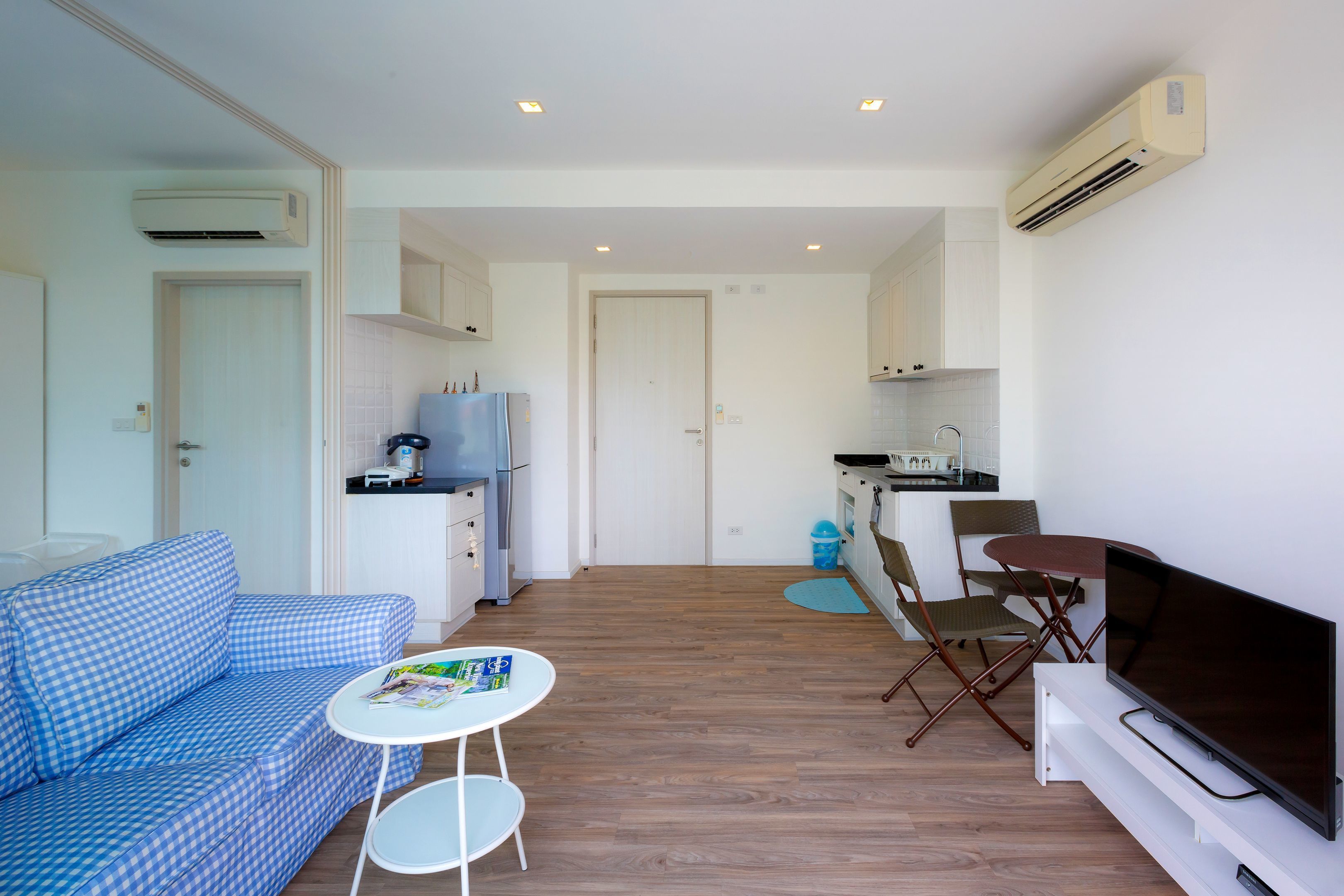 A modern living room with a blue checkered sofa, a small white table, a flat-screen TV, and a kitchen area with cabinets and a refrigerator.