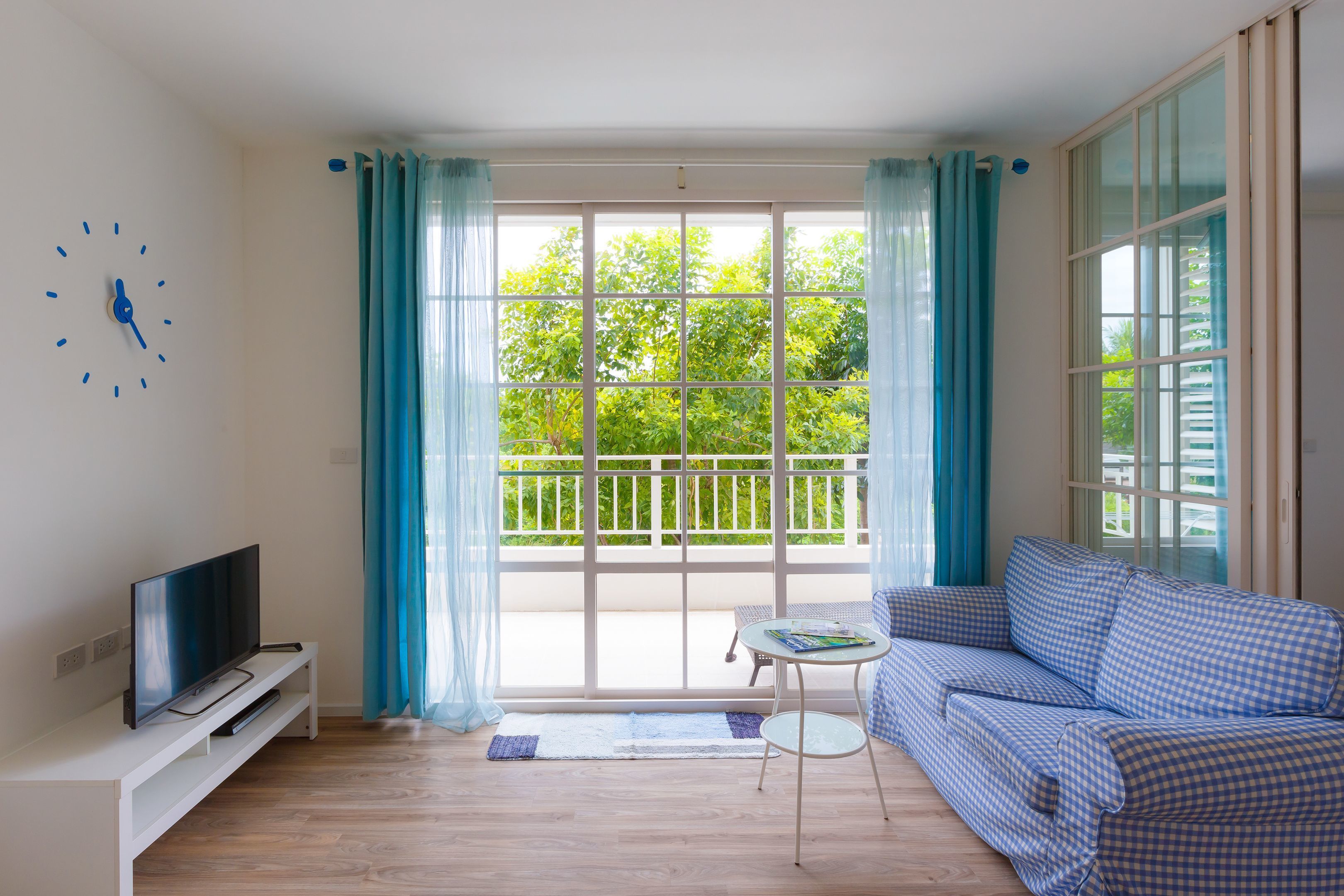 A living room with a television on a white stand, a blue checkered sofa, a small round table, and a large window with curtains.