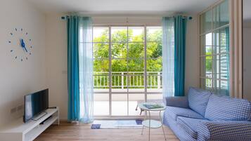 A living room with a television on a white stand, a blue checkered sofa, a small round table, and a large window with curtains.