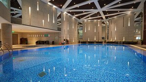 Una amplia piscina interior con suelo de baldosas, rodeada por un techo alto con luminarias lineales, y una zona de recepción con mostrador y sillas.