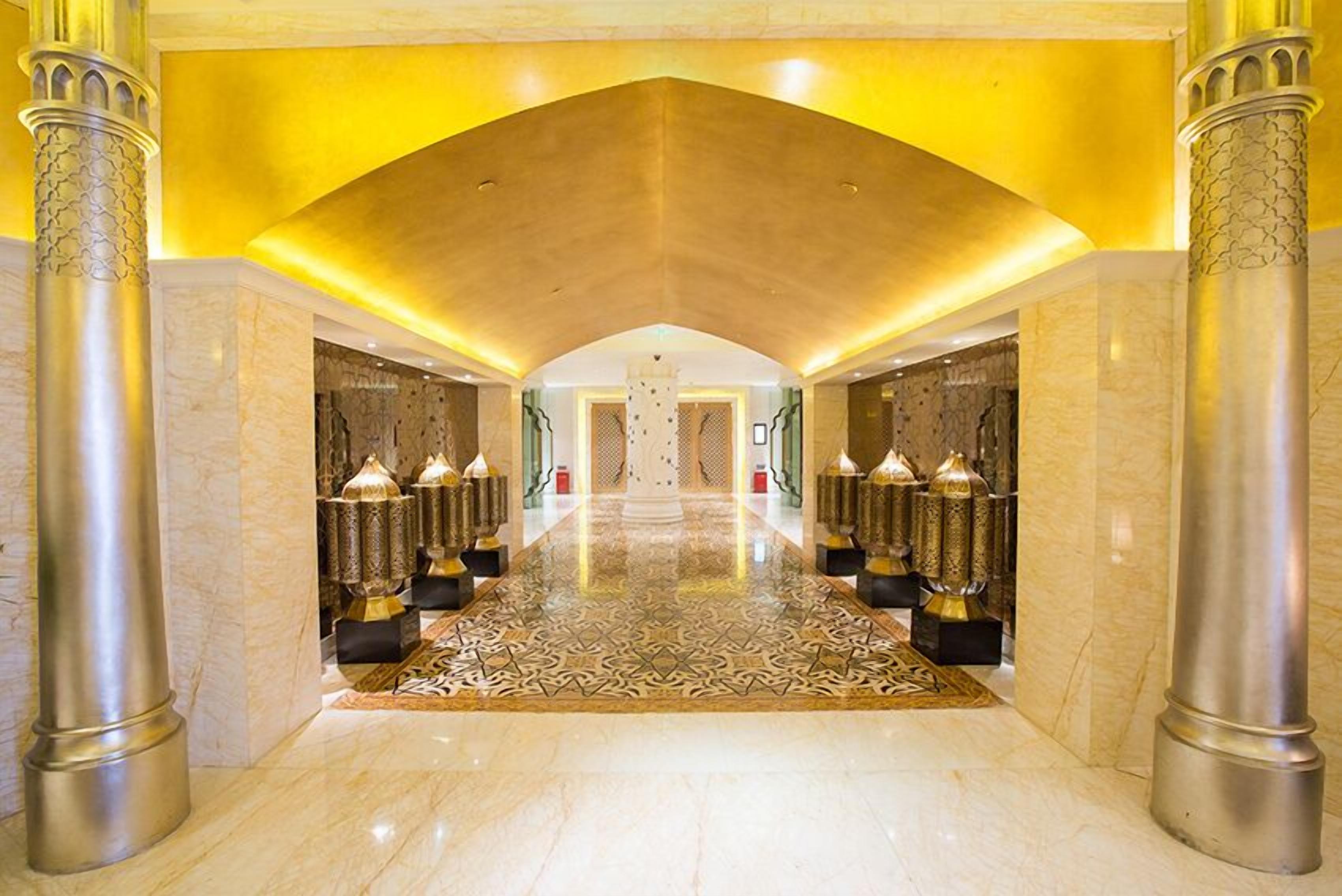 A grand hallway with marble flooring, ornate columns, and a patterned carpet leading to a central area with a chandelier.