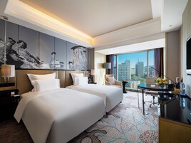 A hotel room with two beds, a desk, a chair, a TV, and a large window with a city view.