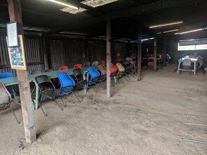 A covered outdoor seating area with multiple tables and chairs, a dirt floor, and a corrugated metal wall. - The Woolshed at Jondaryan - Campsite (Jondaryan)