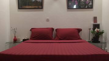 A bed with red bedding, two pillows, a glass table with a vase and a plant, and two framed pictures on the wall.