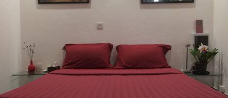 A bed with red bedding, two pillows, a glass table with a vase and a plant, and two framed pictures on the wall.