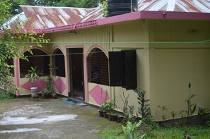 A single-story building with a corrugated metal roof, a pink and beige facade, and a small porch area with potted plants.
