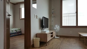 Habitación de hotel con piso de madera, un televisor sobre un mueble de madera, un escritorio, una silla, un ventilador y una ventana con persianas.
