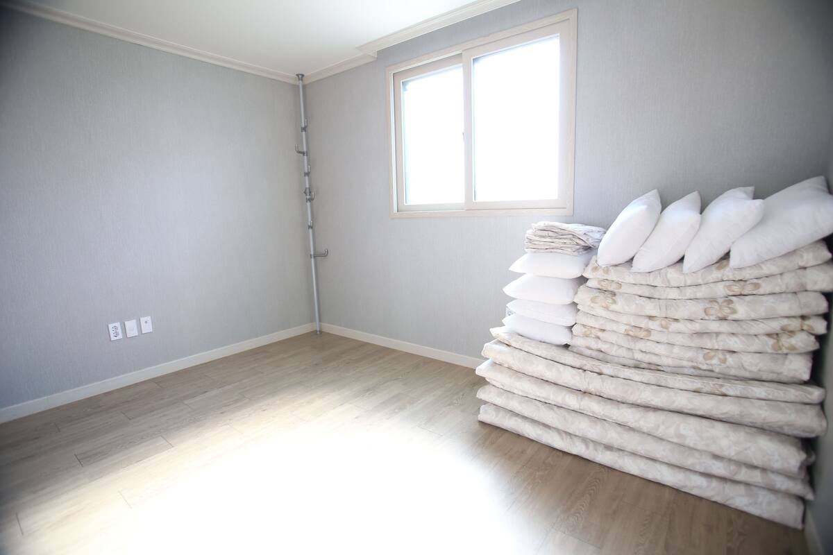 Una habitación con paredes grises claras, suelo de madera, una ventana con persianas y una pila de almohadas.