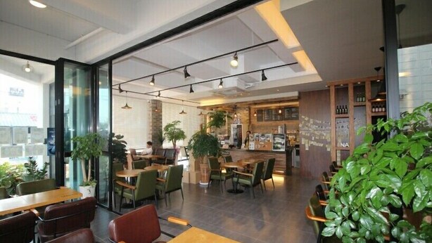 Un café moderno con mesas y sillas de madera, una zona de bar y plantas de interior.