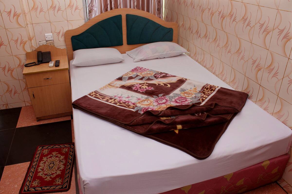 A neatly made bed with a floral bedspread, a wooden nightstand with a phone and remote, and a red patterned rug on the floor.