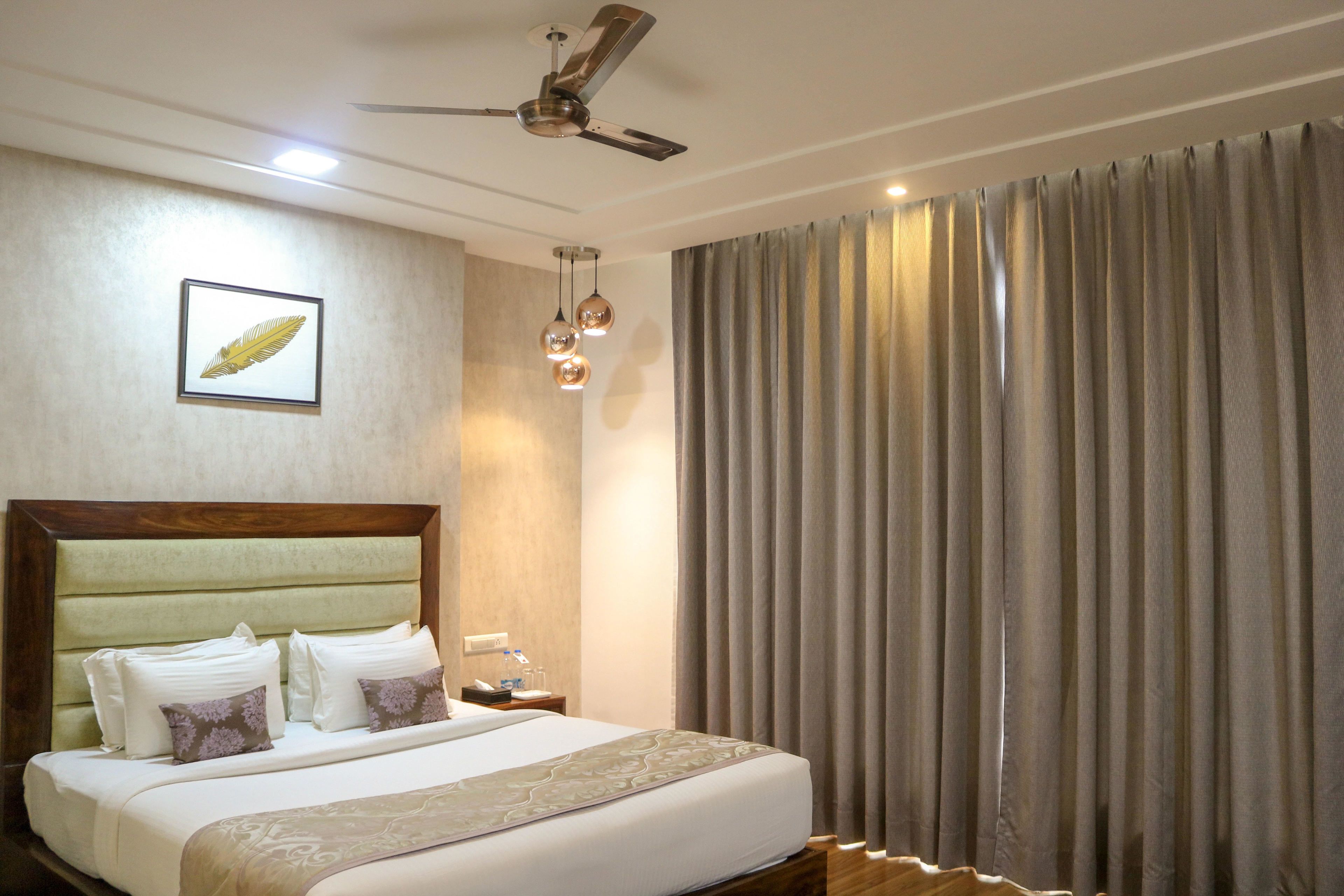 Una habitación de hotel con una cama grande, un ventilador de techo, una obra de arte enmarcada y un ventanal con cortinas.