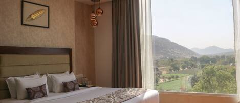 A hotel room with a large bed, a headboard, a framed picture on the wall, and a view of a mountainous landscape through a window.