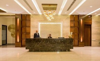 A reception desk with two individuals standing behind it, a patterned wall in the background, and a chandelier above.