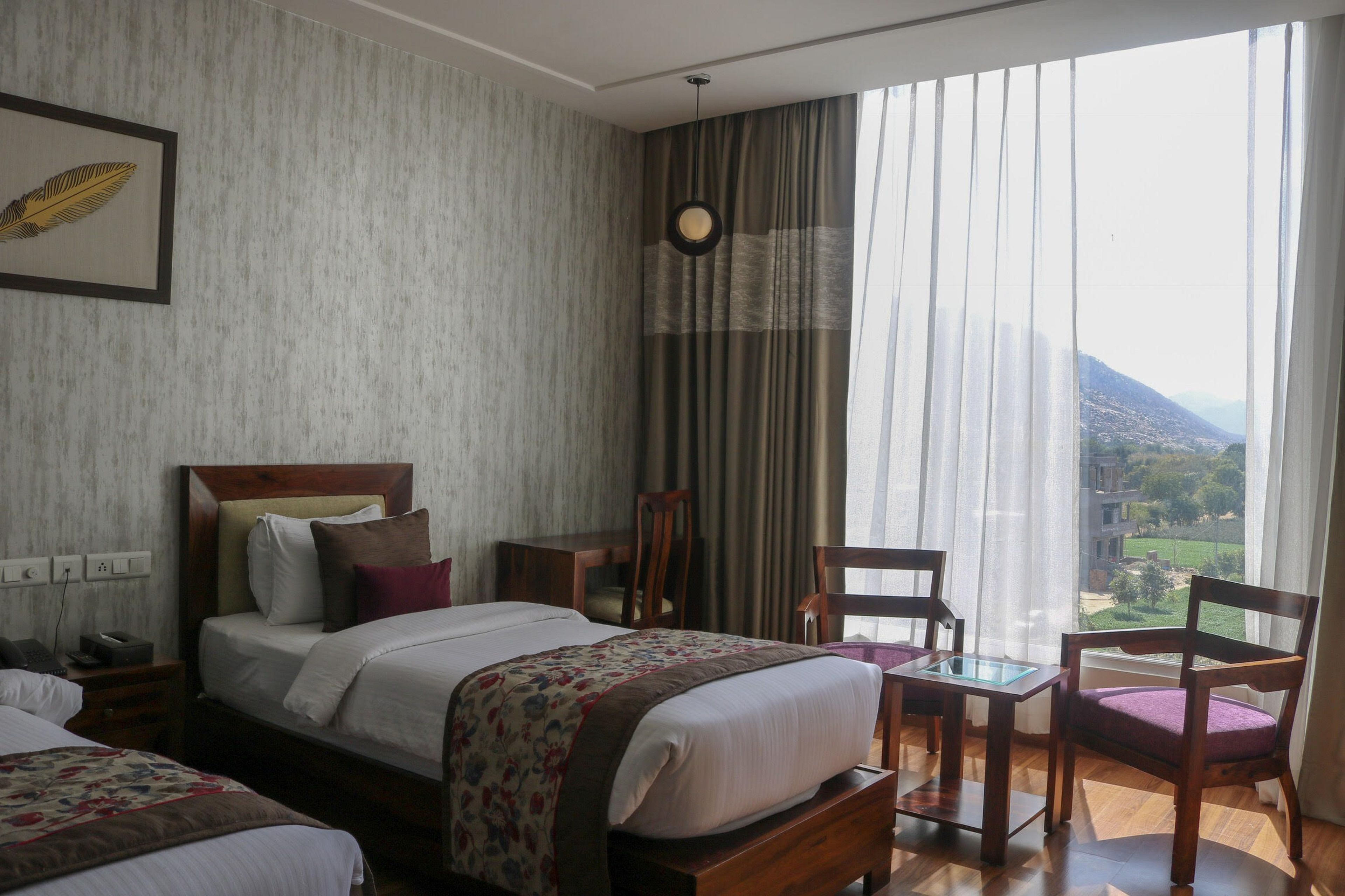 A hotel room with a large bed, two chairs, a small table, and a view of a mountainous landscape through sheer curtains.