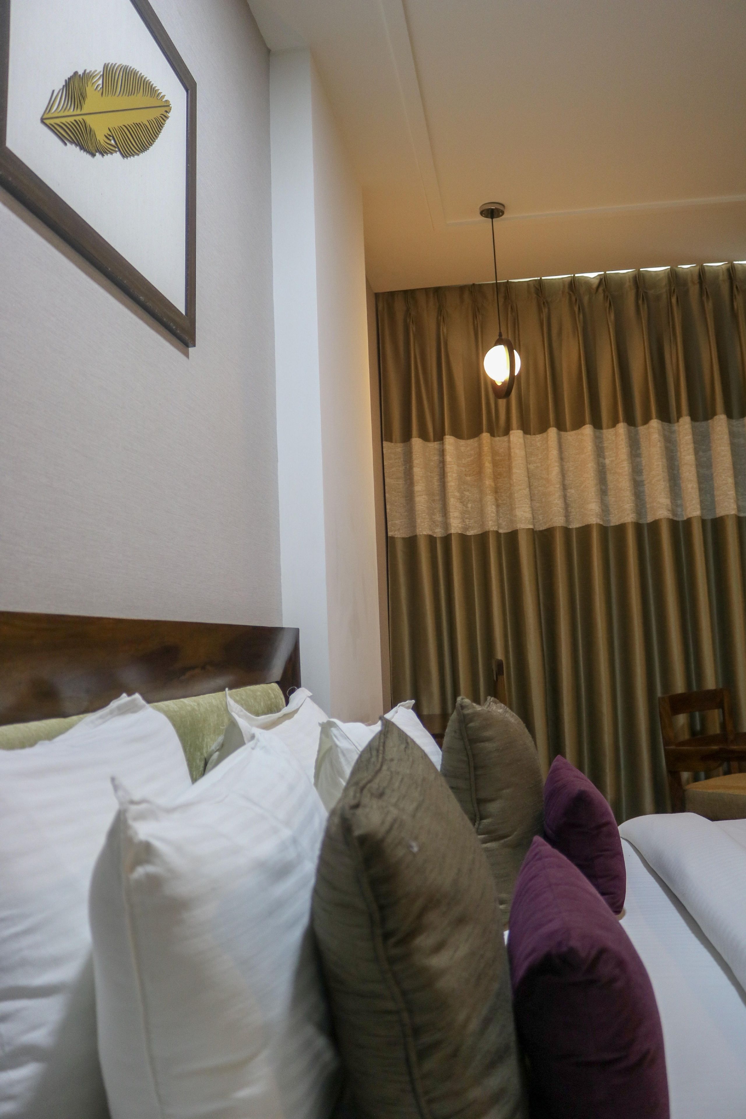 A hotel room with a bed, pillows, a headboard, a hanging light fixture, and a window with curtains.