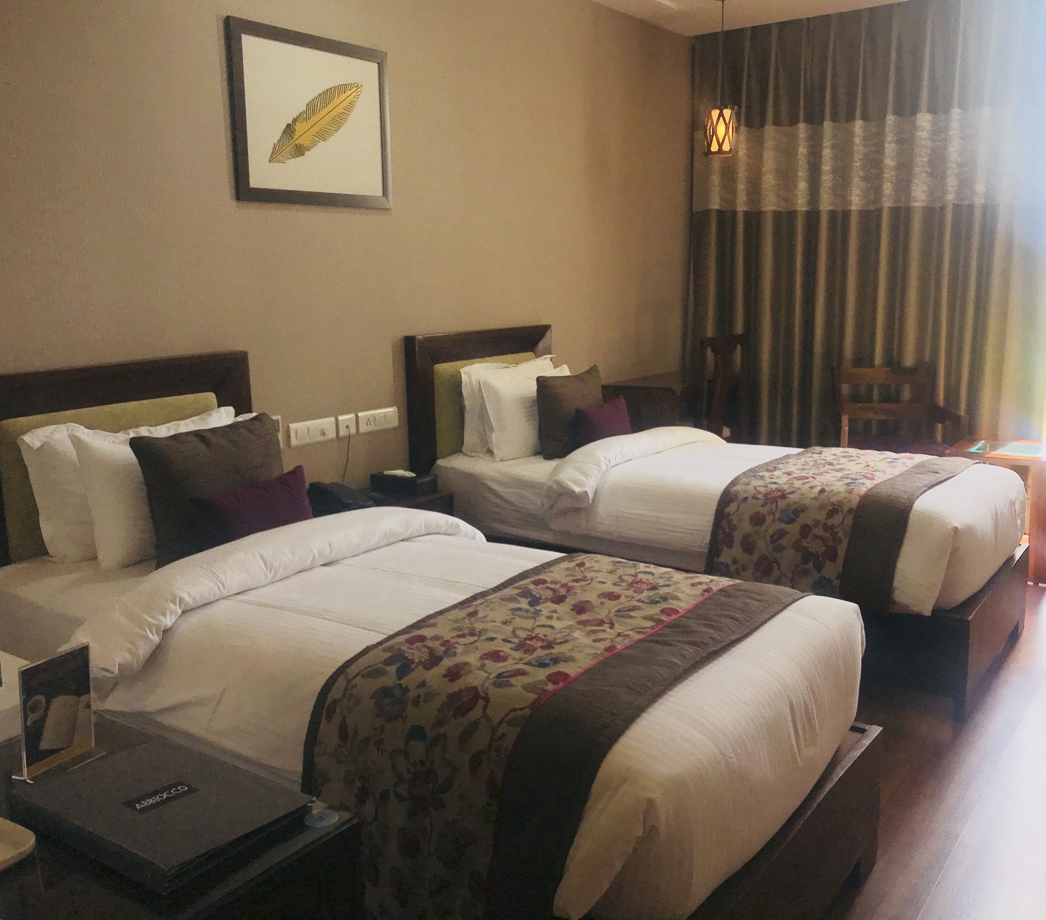 Habitación de hotel con dos camas, cada una con ropa de cama blanca y colchas con estampado floral. Hay una obra de arte enmarcada en la pared sobre las camas y una lámpara colgante.