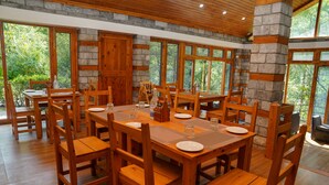 Un comedor con mesas y sillas de madera, detalles de piedra en las paredes y amplias ventanas con vistas a la vegetación.