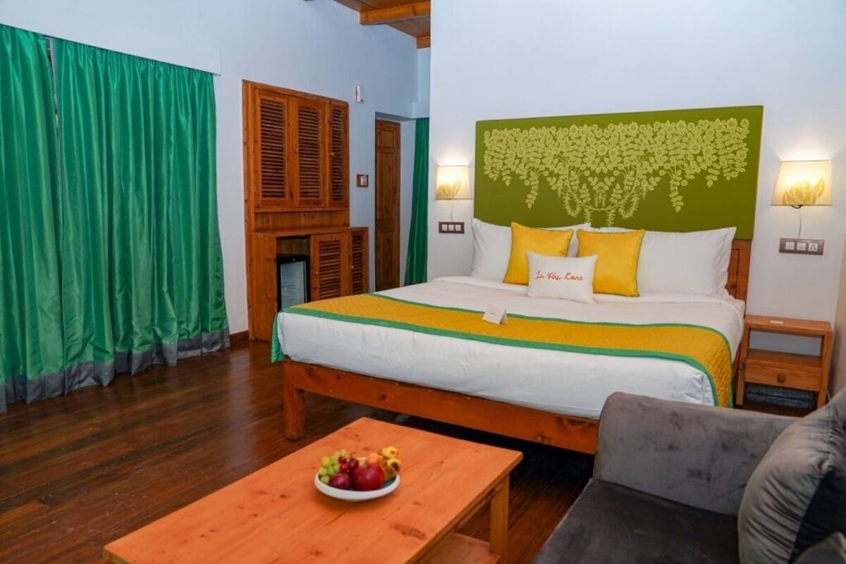 A bedroom with a bed, green curtains, a wooden headboard, a bedside table, and a fruit bowl on a table.