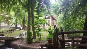 Un puente de madera rústico sobre un arroyo pequeño, rodeado de vegetación exuberante y un edificio de piedra al fondo.