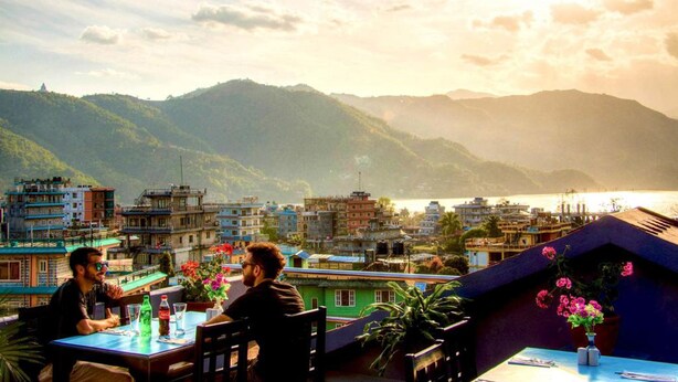 A terrace with a table and chairs, overlooking a mountainous landscape with buildings and a body of water. - Hotel Atrium (Pokhara)