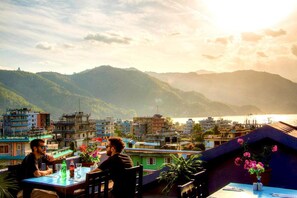 A terrace with a table and chairs, overlooking a mountainous landscape with buildings and a body of water. - Hotel Atrium (Pokhara)