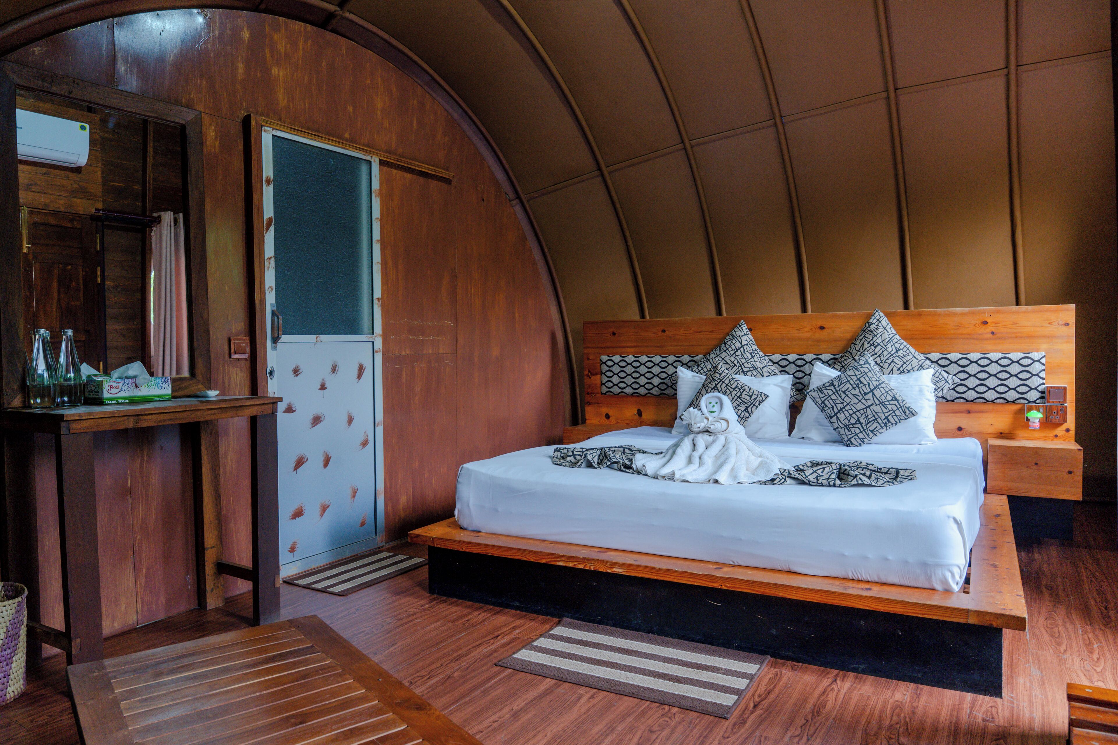 Quarto com cabeceira de madeira, cama com roupa de cama branca e almofadas estampadas. Tem uma mesa de cabeceira de madeira com uma luminária e uma pequena mesa com uma garrafa e copos.