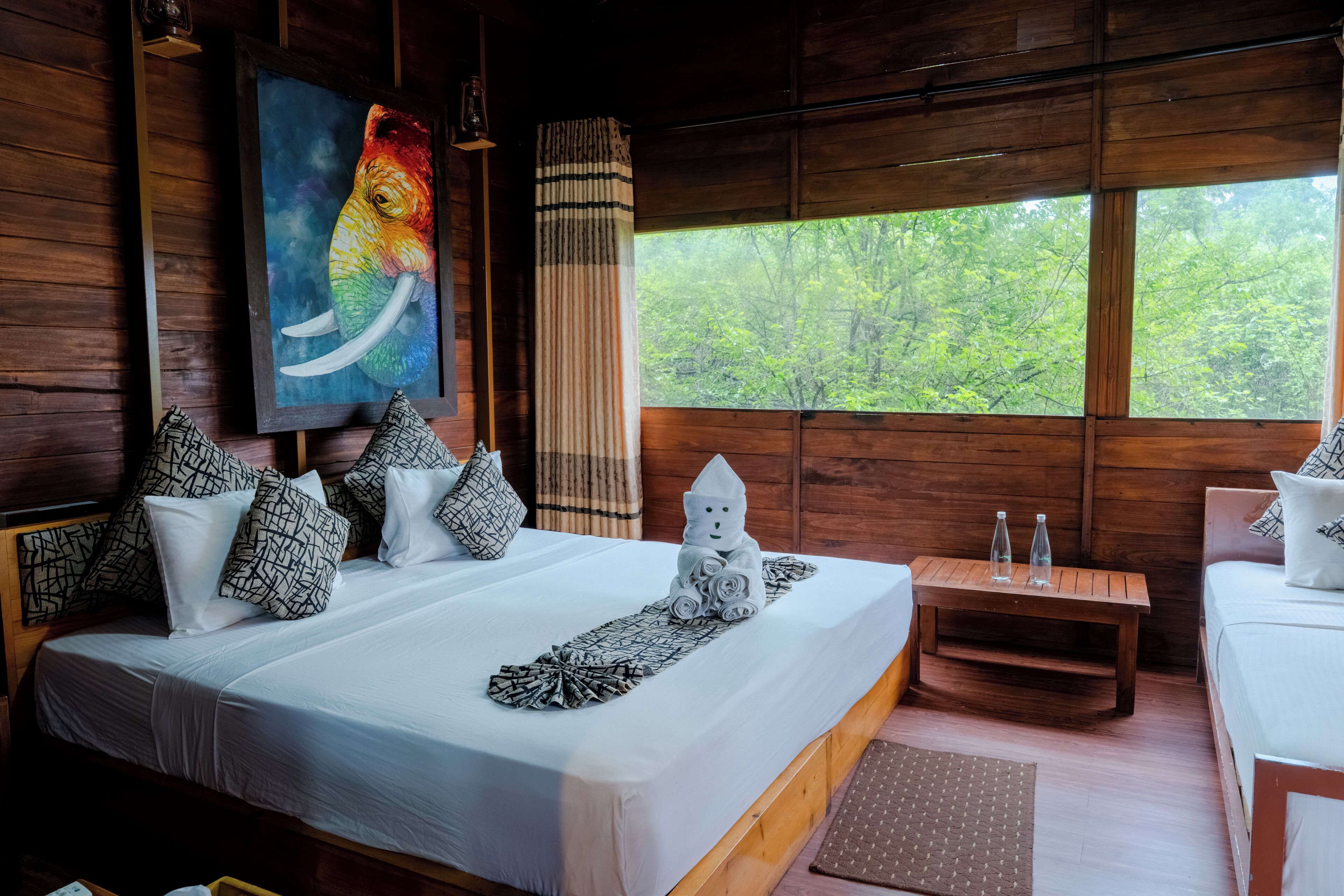 A wooden cabin bedroom with a large bed, two smaller beds, a painting of an elephant, and a view of trees through the window.