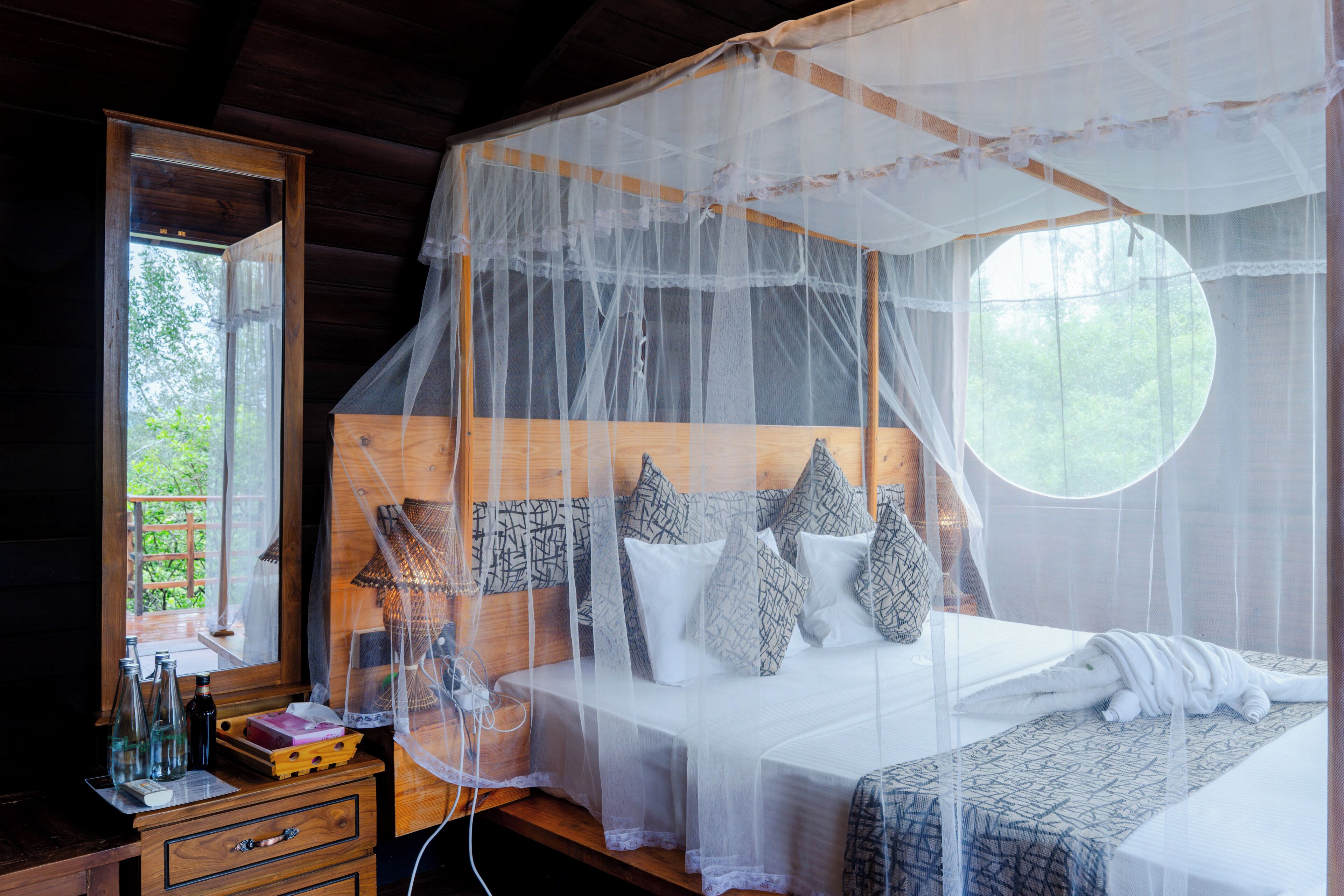 A bedroom with a canopy bed, a wooden dresser, and a window with a view of greenery.