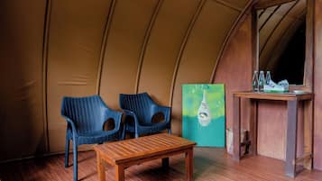 Um quarto com tenda, duas cadeiras azuis, uma mesa de madeira, um quadro e uma pequena mesa de madeira com garrafas.