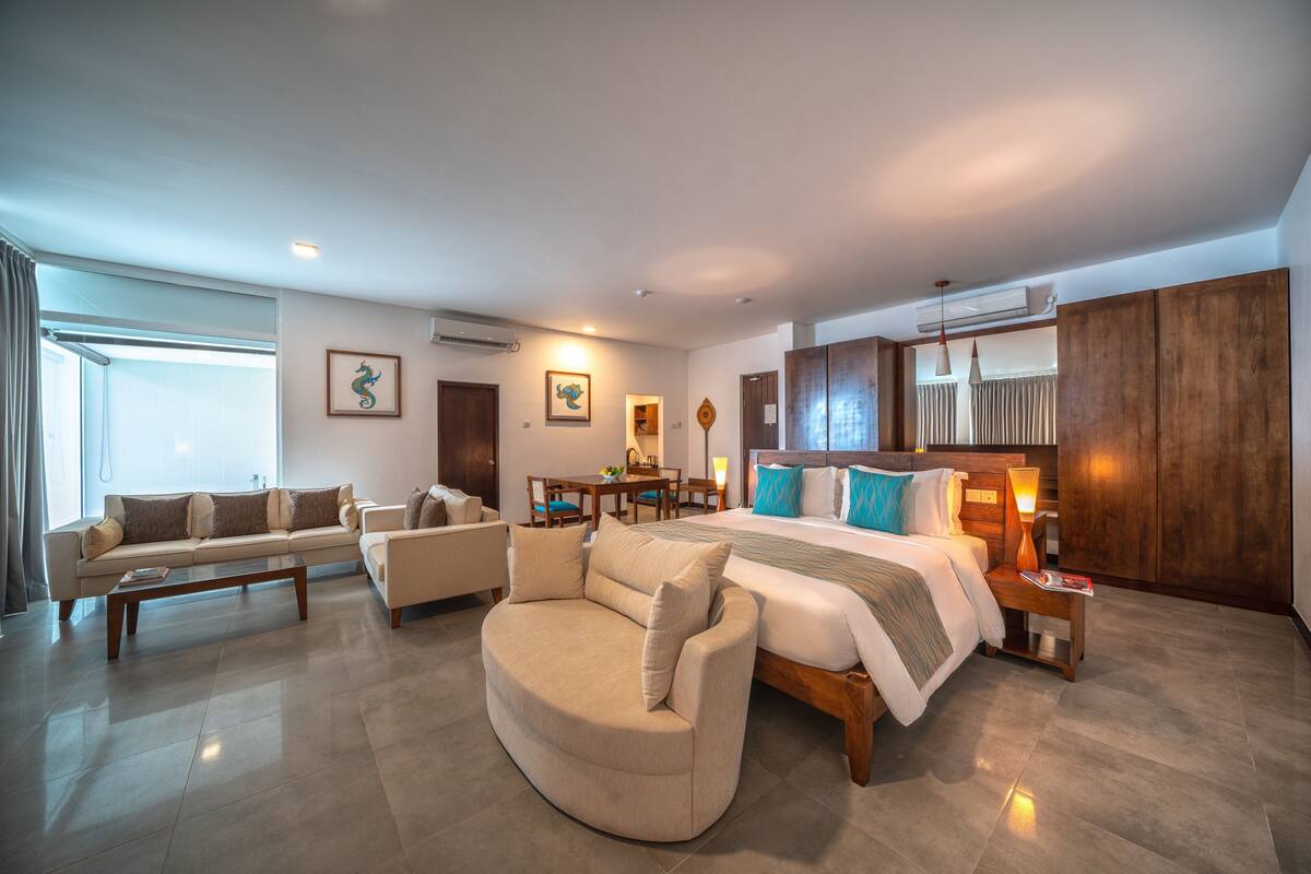 A modern hotel room with a large bed, a sofa, a curved armchair, and a dining area.