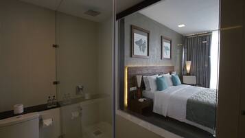 Quarto de hotel moderno com box de vidro, cama bem arrumada com cabeceira de madeira e quadros nas paredes.