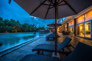 Zona de piscina con sillas de descanso y sombrillas, piscina y un edificio con amplias ventanas.