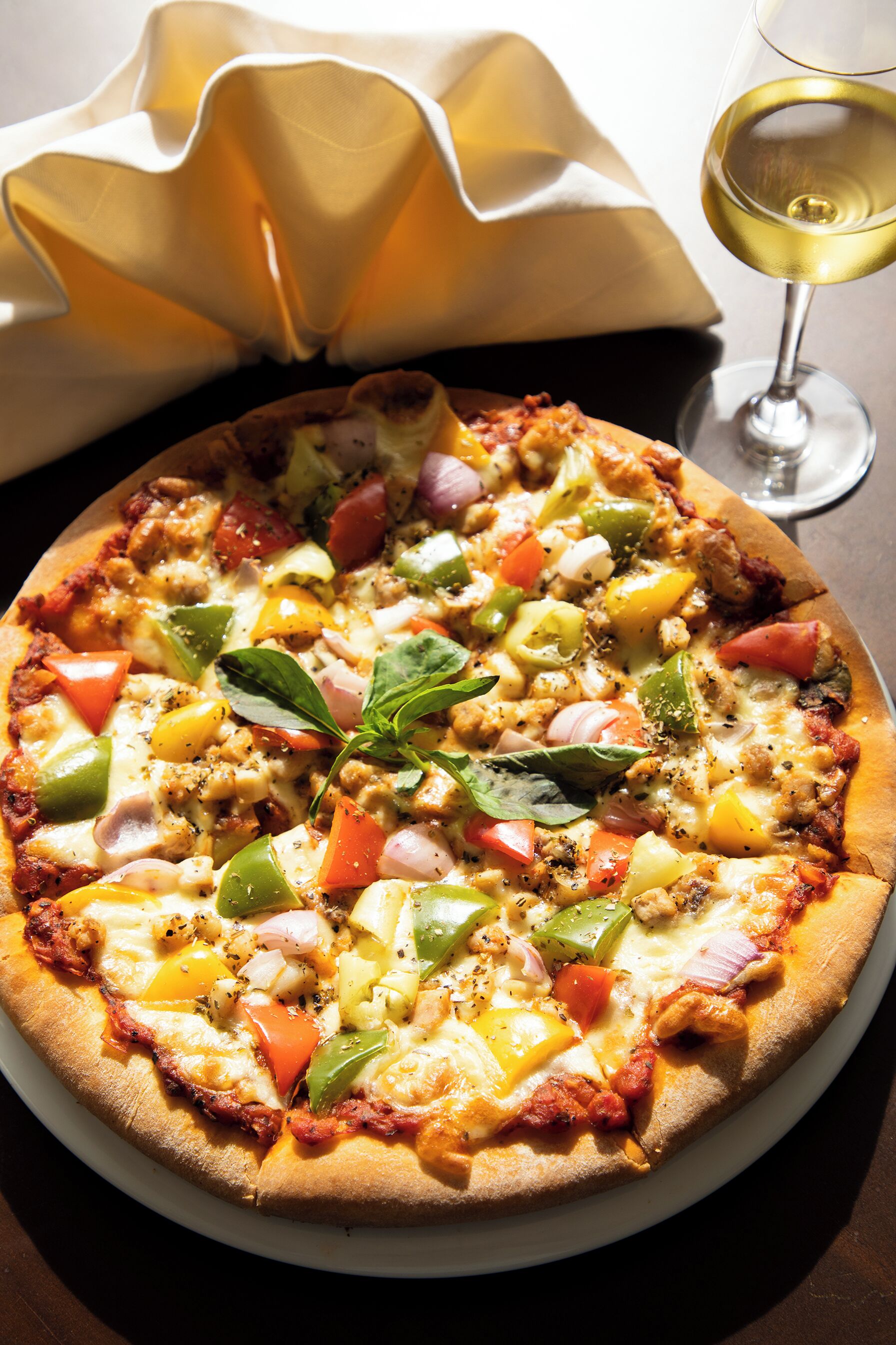 A pizza with various toppings on a white plate, accompanied by a glass of white wine and a folded napkin.