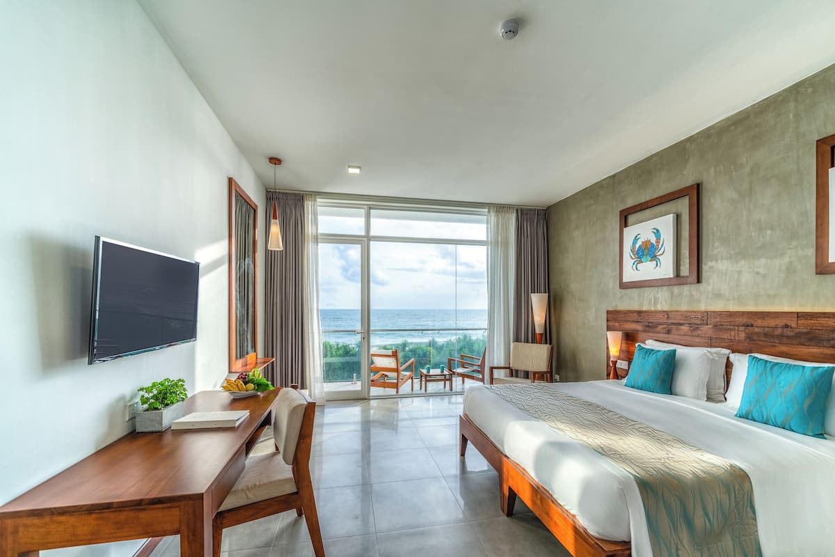 A hotel room with a bed, a desk, a TV, a large window with a view of the ocean, and a ceiling fan.