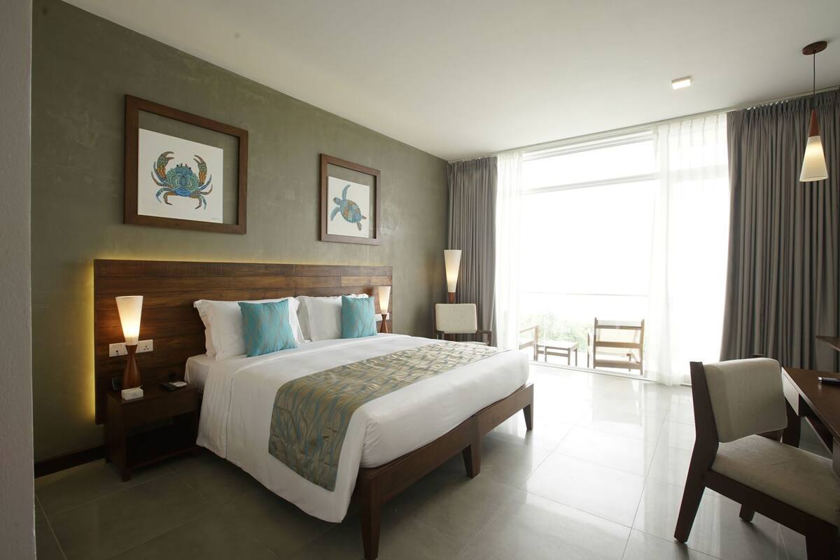 A hotel room with a large bed, two nightstands, a desk with a chair, a balcony with a view, and artwork on the walls.