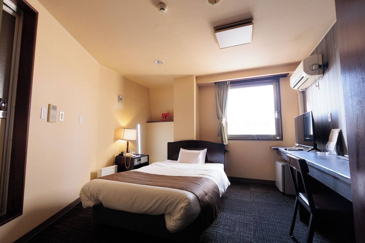 A hotel room with a bed, desk, chair, lamp, and a window.
