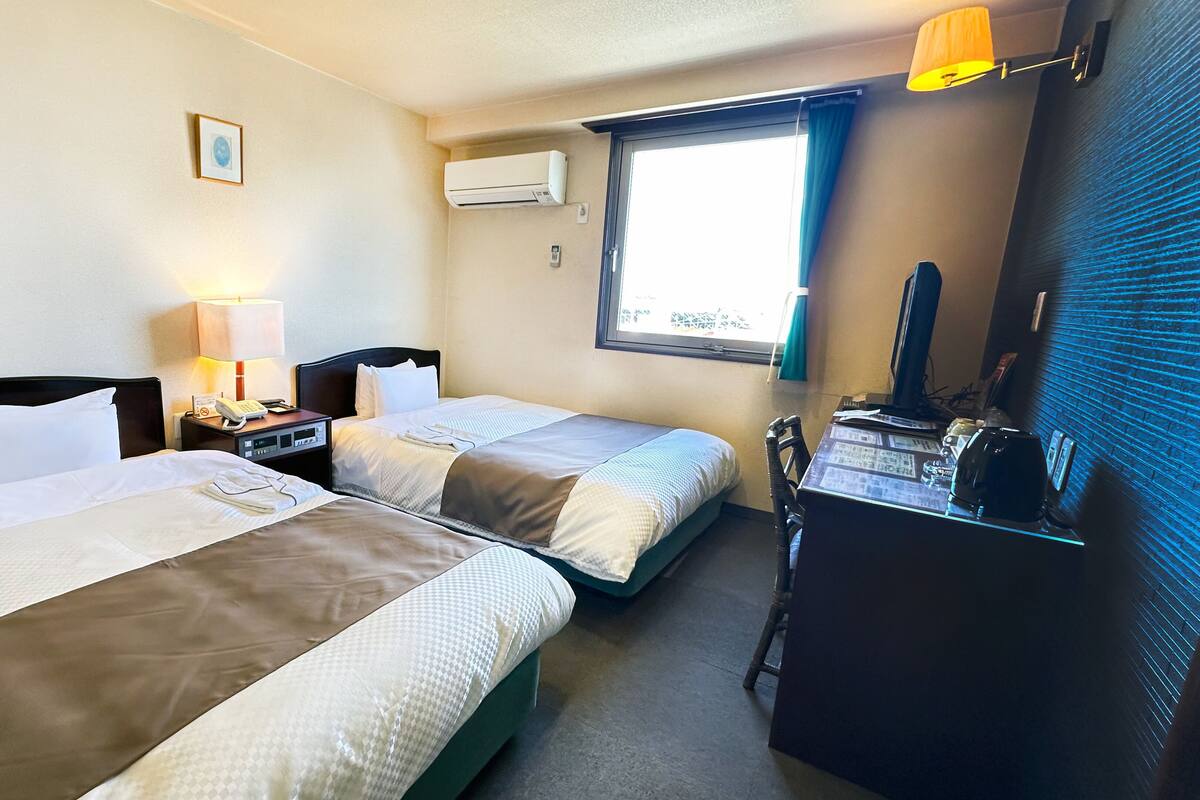 A hotel room with two beds, a desk with a computer, an air conditioner, and a window with curtains.