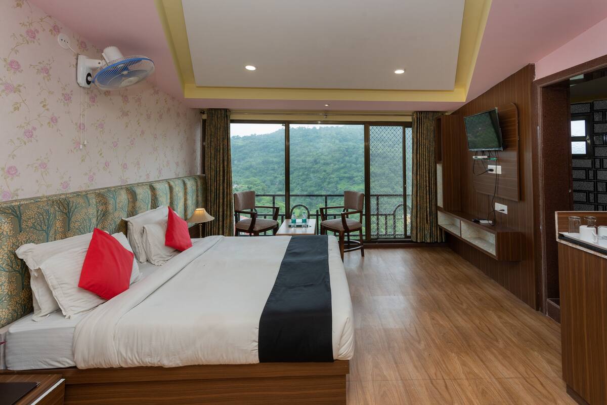 Een hotelkamer met een groot bed, een balkon met uitzicht op de bergen, een aan de muur bevestigde televisie en een kleine tafel met stoelen.