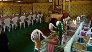 A tented dining area with tables set for multiple people, white plastic chairs, and a central table with a red and white checkered tablecloth. - Nomad Camp (Leh)