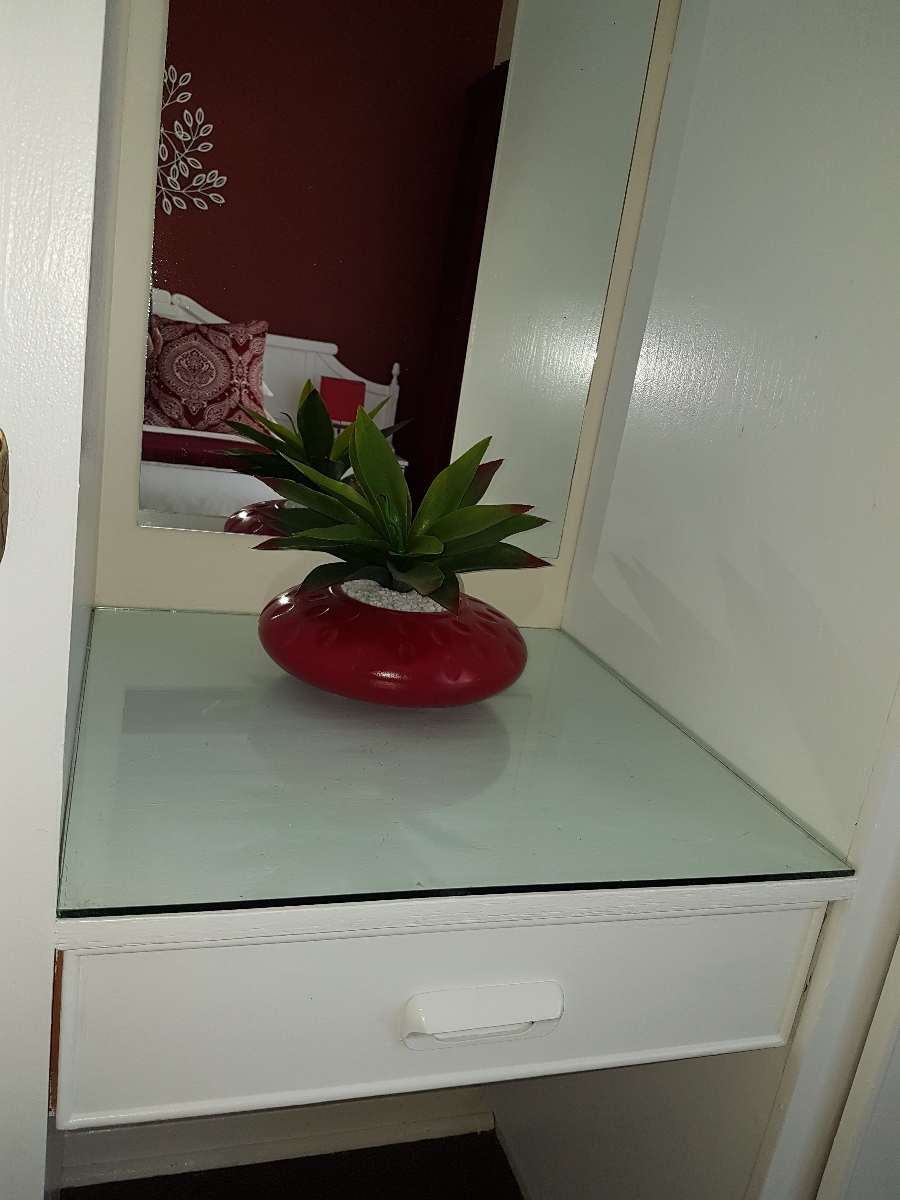 Un petit meuble blanc, doté d’un dessus en verre et contenant un seul pot rouge dans lequel se trouve une plante verte.