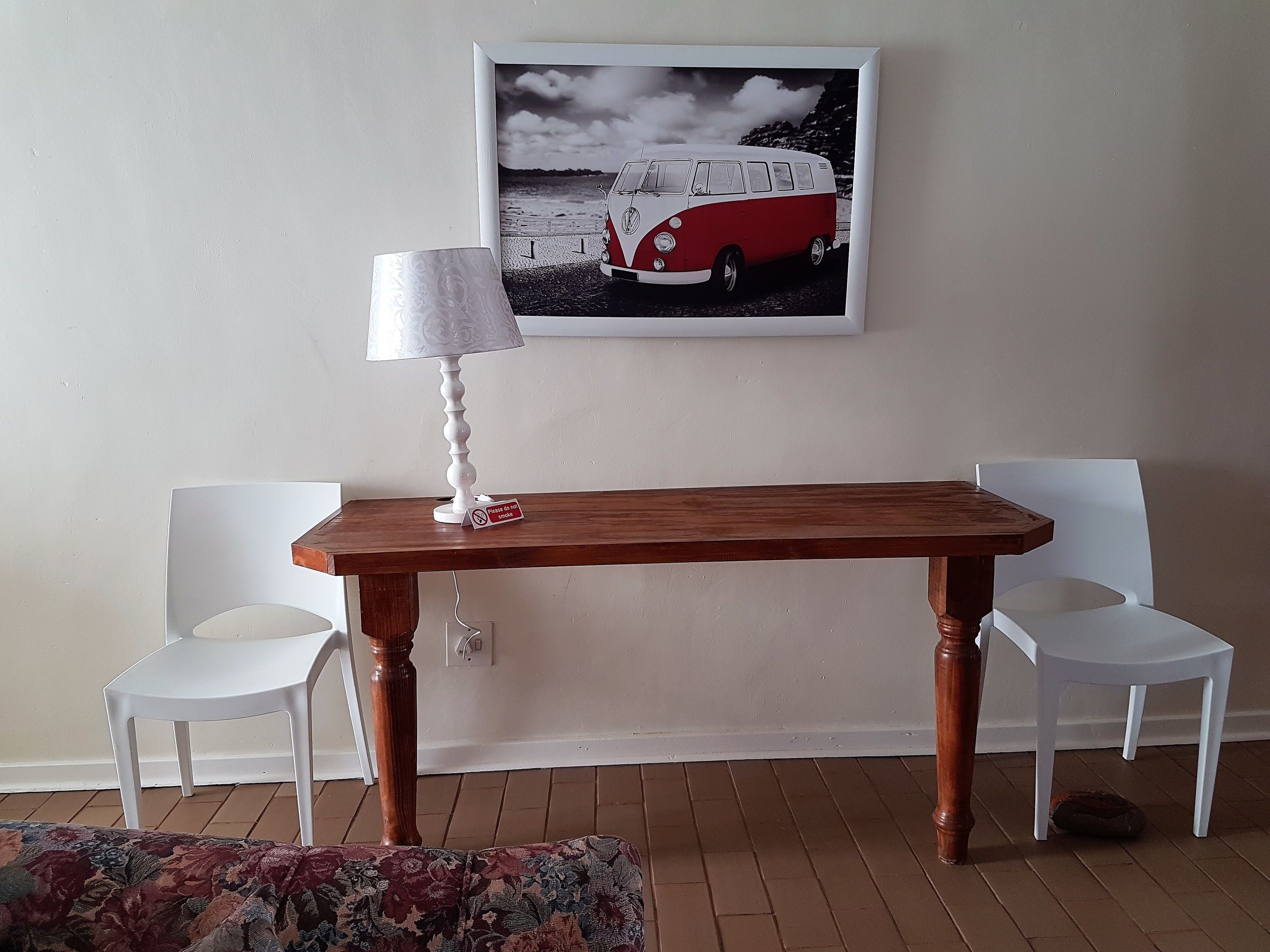 Una habitació amb una taula de fusta, dues cadires blanques, una làmpada i un quadre al mur.