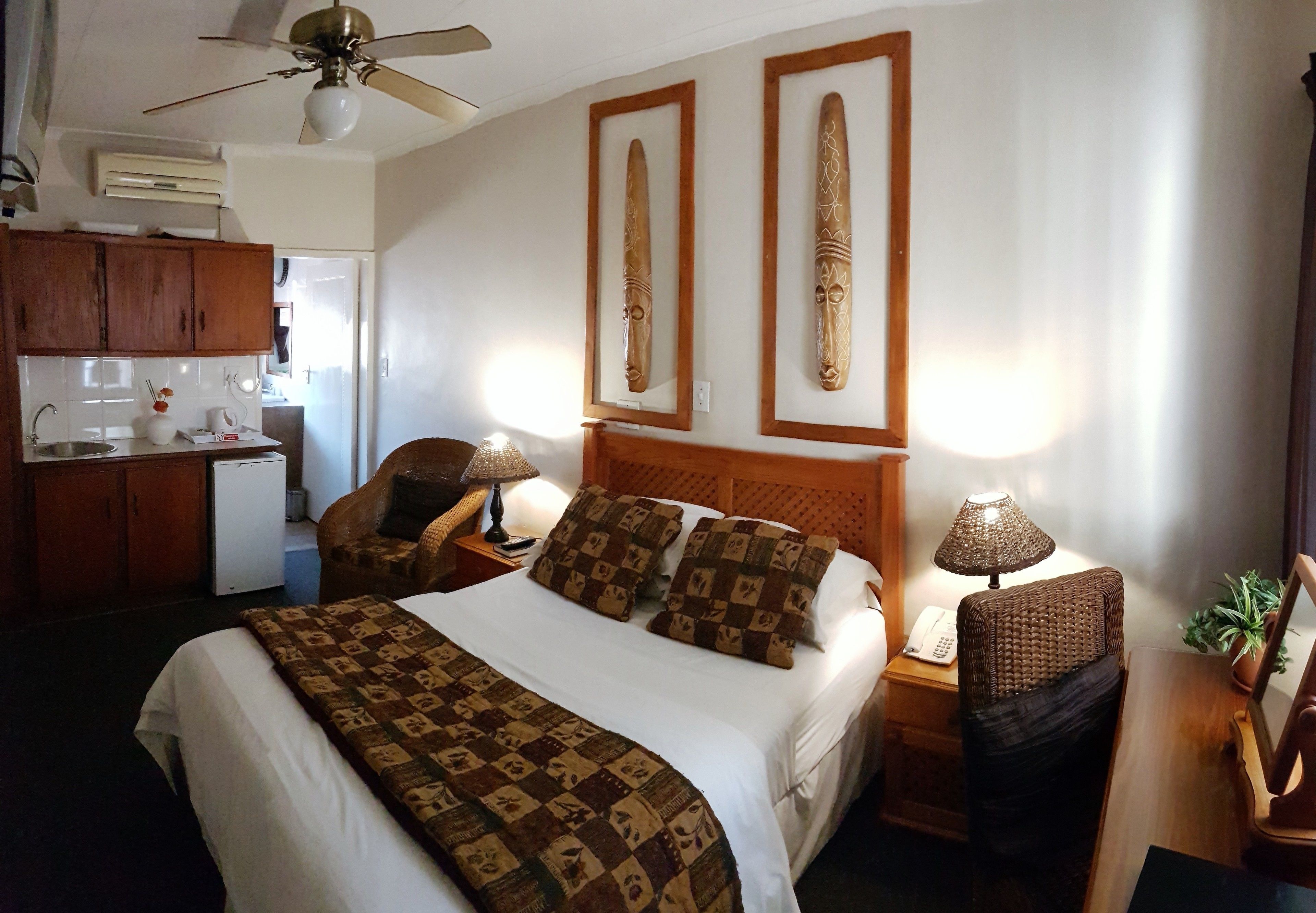 A hotel room with a bed, two wall-mounted headboards, a desk with a lamp, a chair, and a small kitchenette.