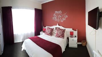 Habitación de hotel con una cama grande, cabecera blanca, almohadas con estampado rojo y blanco, una lámpara de noche roja, una mesita de noche blanca, un televisor de pared y una ventana con cortinas.