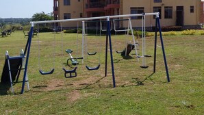 Un parc infantil amb cadira de balanç i tobogan davant d'un edifici de dues plantes.
