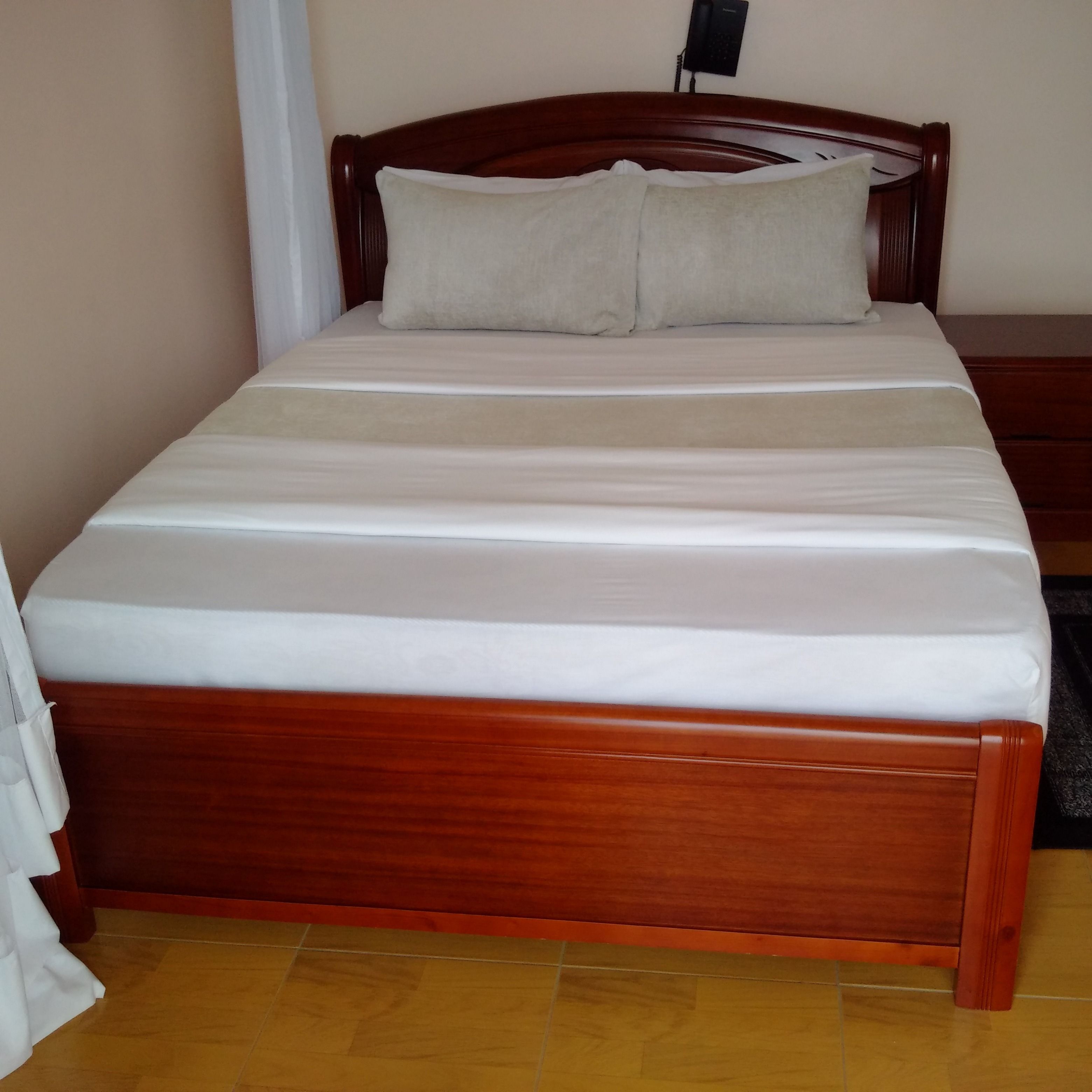 Ein hölzernes Bett mit weißer Matratze und zwei Kissen, ein Nachttisch mit Telefon und ein Fliesenboden.