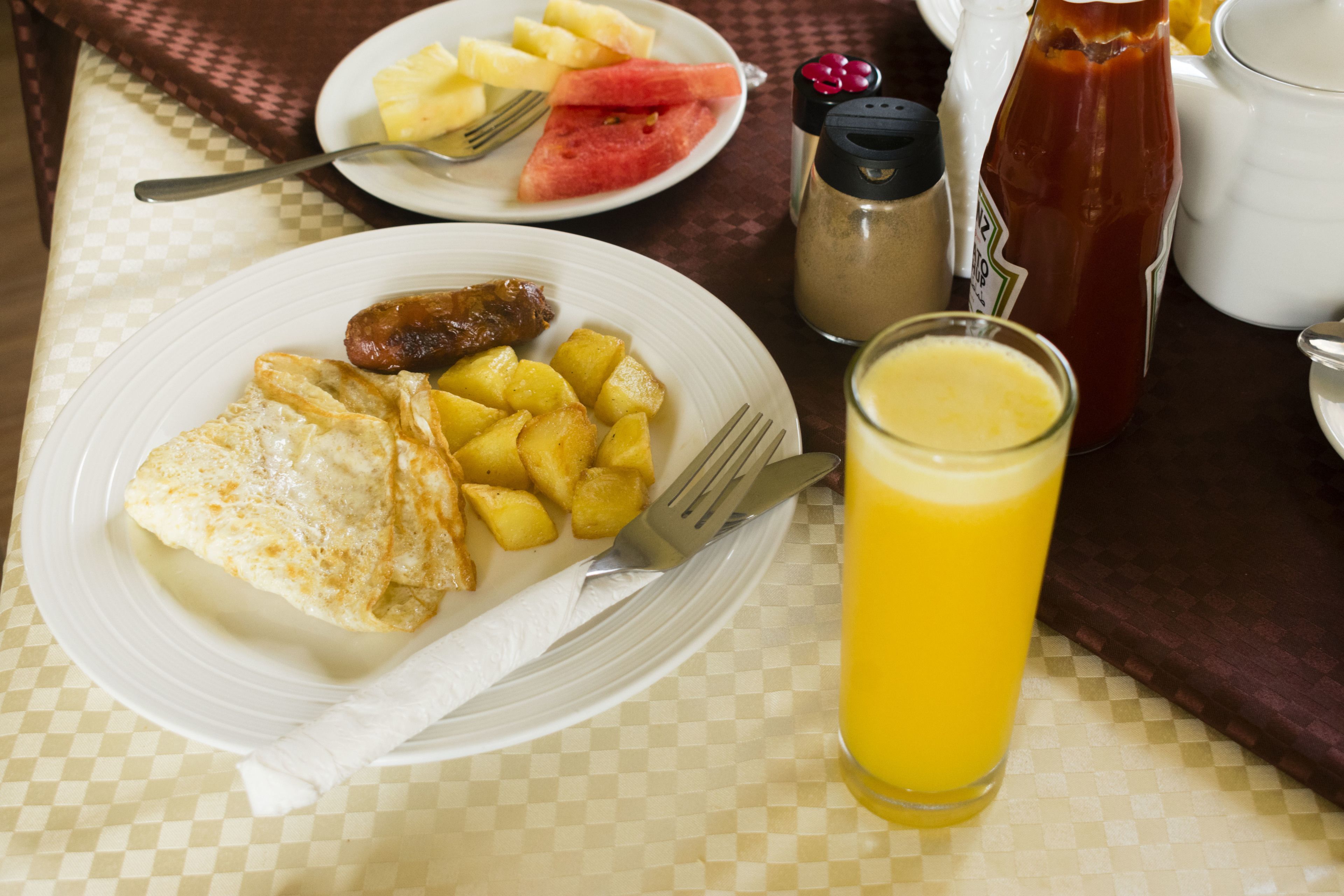 Une assiette de petit-déjeuner avec des œufs, des pommes de terre, des saucisses et un verre de jus d’orange, posée sur une table recouverte d’une nappe à carreaux.