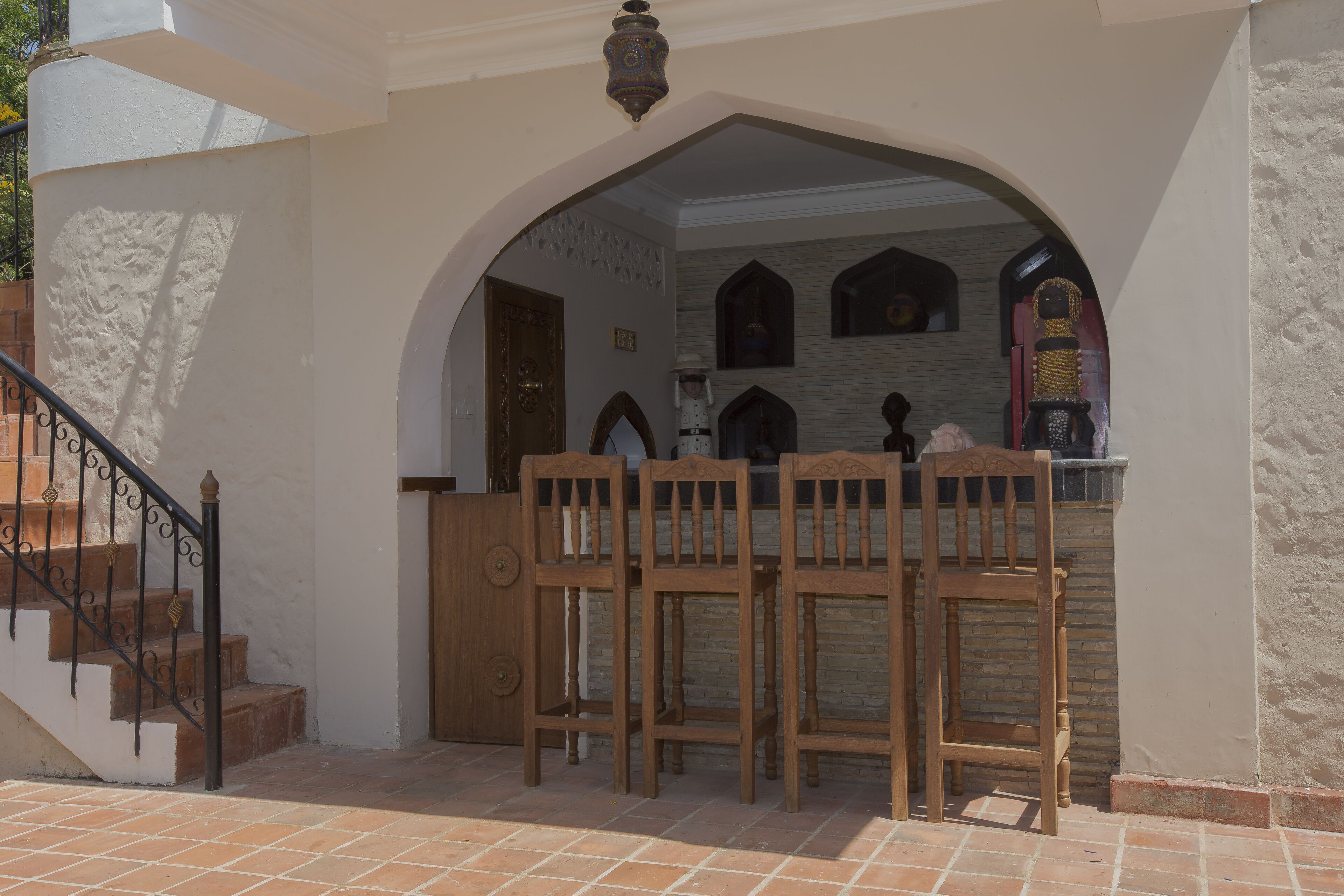 Un área de estar al aire libre con techo, sillas de madera y una mesa, una escalera con barandilla de metal y una lámpara decorativa en el techo.
