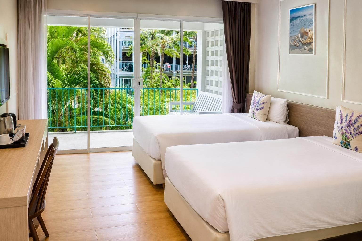 A hotel room with two beds, a desk with a coffee maker, and a view of a balcony with greenery.