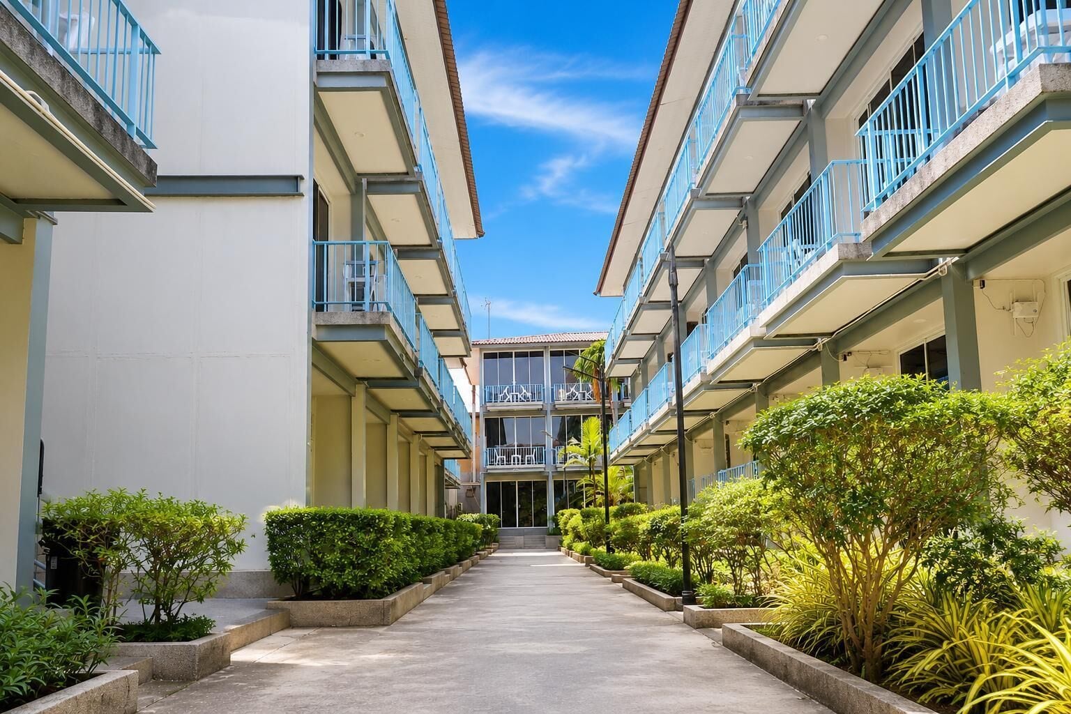 A hotel corridor with multiple balconies, a central staircase, and greenery along the sides.