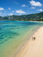 A beach with clear turquoise water, a sandy shore, and a hilly background with buildings.
